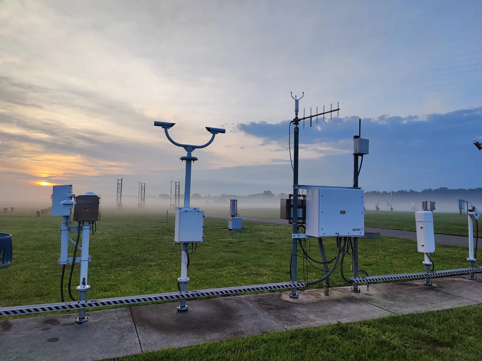 ASOS weather instruments on metal posts in a grassy observing area at sunrise, with low mist in the background.